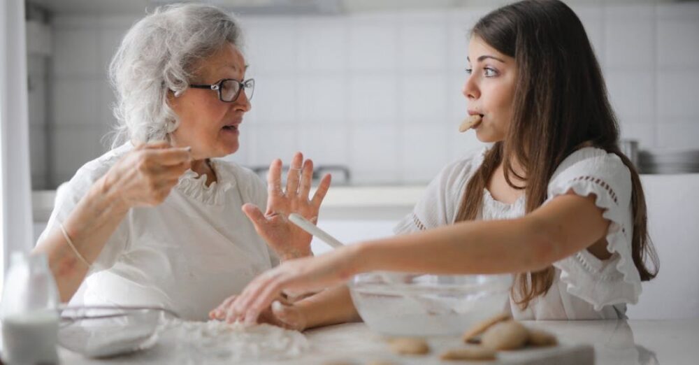 Persuasive Opinions - Calm senior woman and teenage girl in casual clothes looking at each other and talking while eating cookies and cooking pastry in contemporary kitchen at home