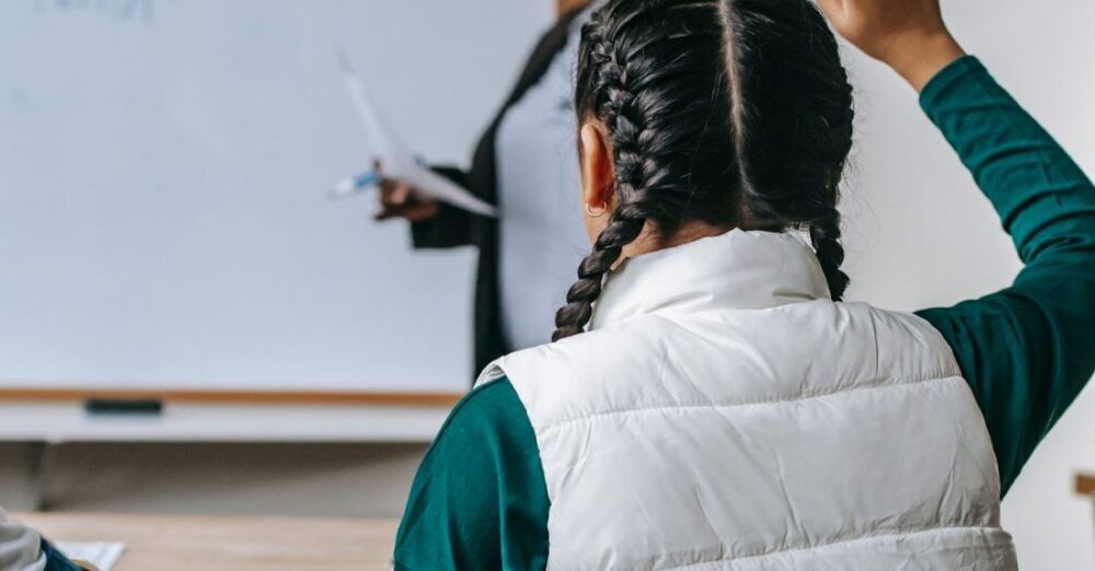 Teacher Training - Smiling African American female teacher standing near whiteboard and looking at schoolgirl raising hand